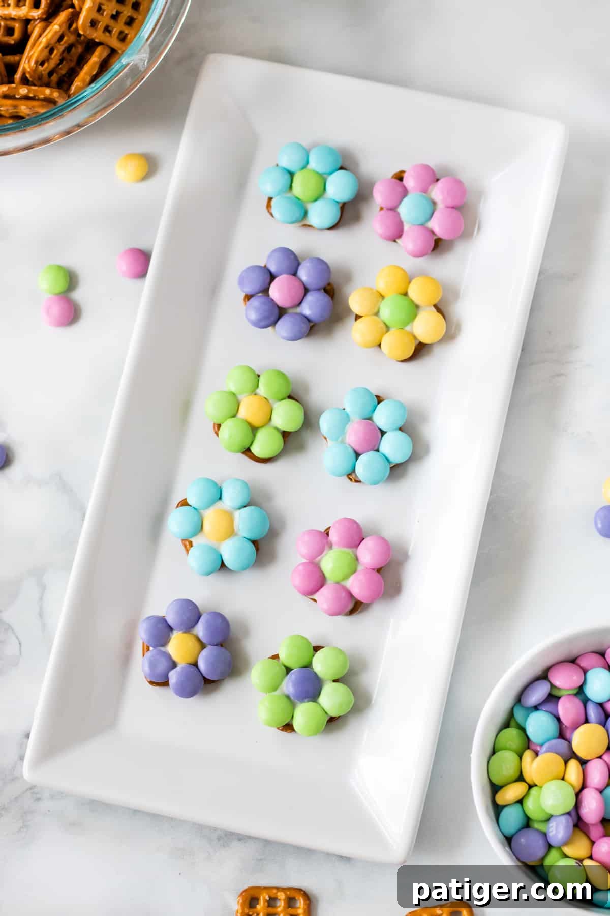 10 pastel-colored flowers made from M&Ms and pretzels on a white serving platter. A bowl of pretzel snaps and a bowl of M&Ms are next to the platter.