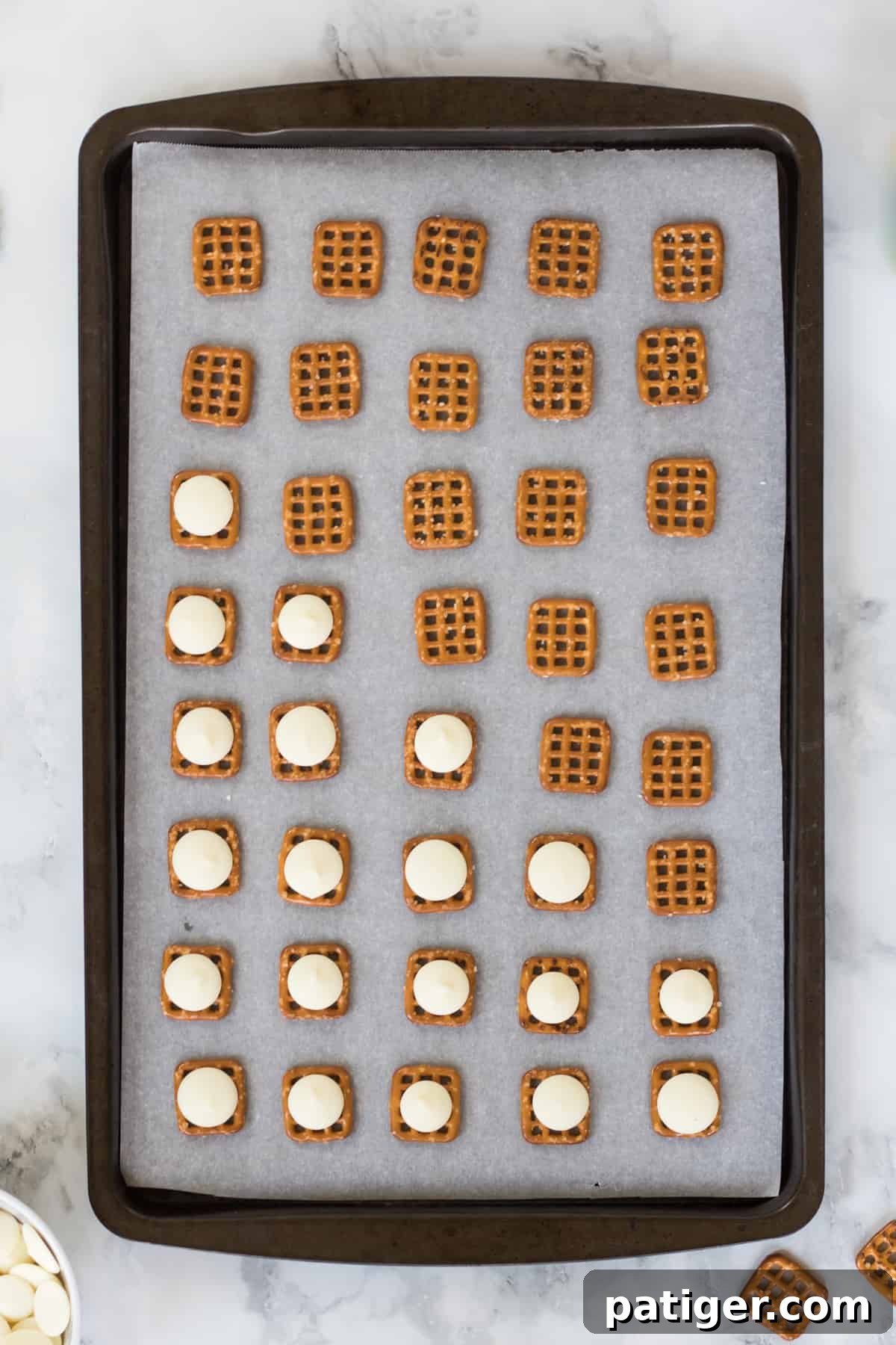 Baking sheet with rows of square pretzels. Half of the pretzels are topped with white chocolate melting wafers.