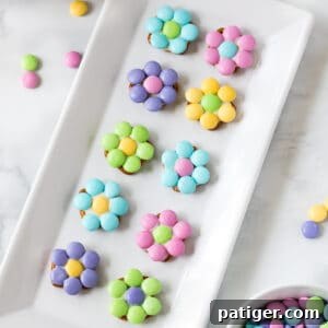 10 pastel-colored flowers made from M&Ms and pretzels displayed on a white serving plate with additional M&Ms sprinkled around the tabletop