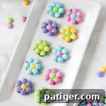 10 pastel-colored flowers made from M&Ms and pretzels displayed on a white serving plate with additional M&Ms sprinkled around the tabletop