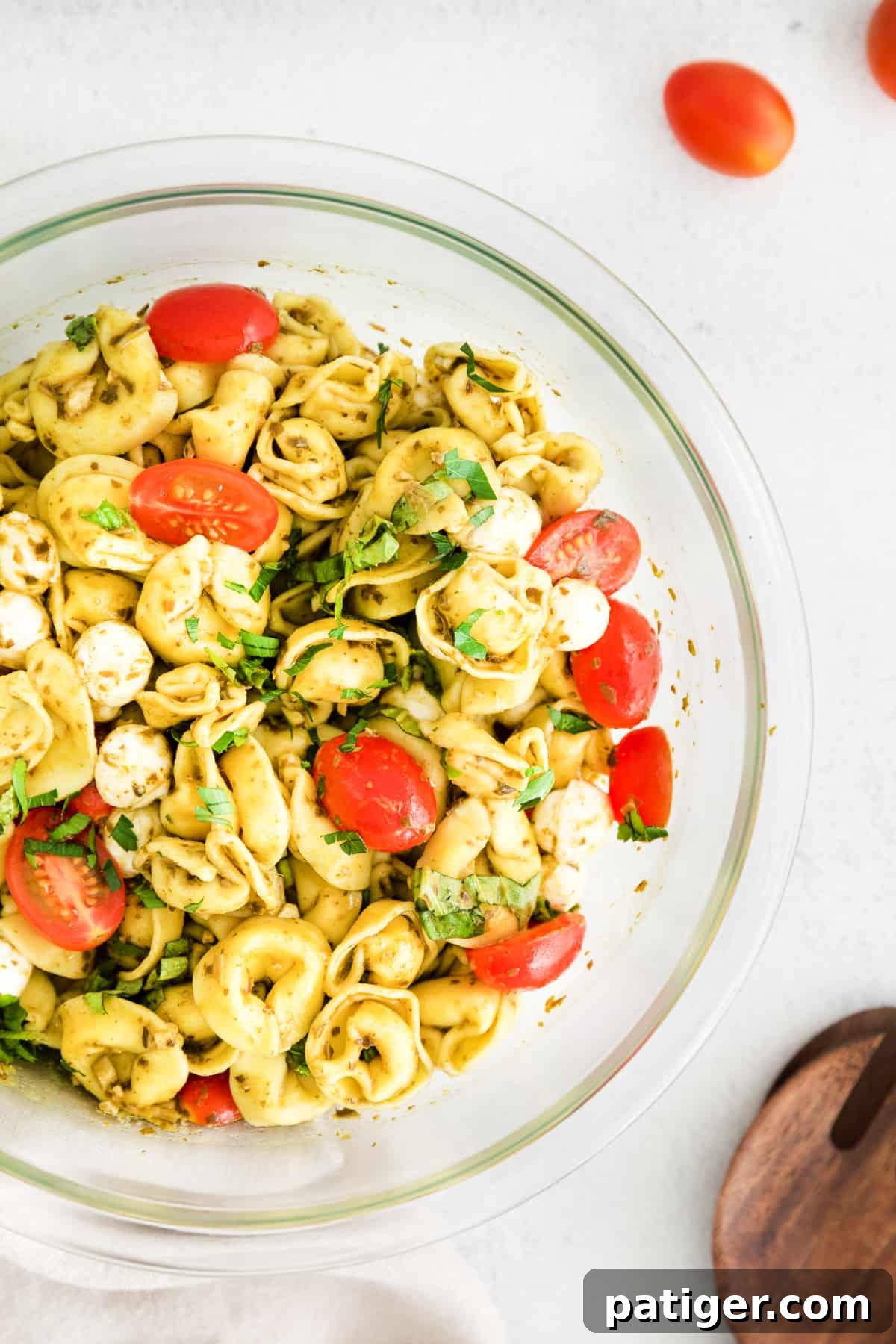 A large glass bowl filled with vibrant tortellini pesto pasta salad, featuring plump cheese tortellini, glistening mozzarella balls, halved grape tomatoes, and fresh basil leaves, ready to be served.