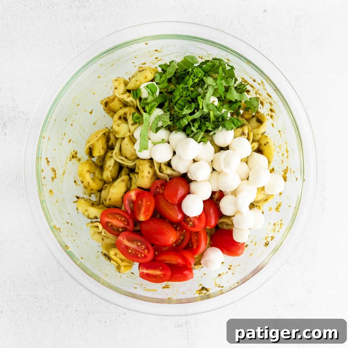A large glass mixing bowl filled with partially assembled pesto pasta salad, showing cooked tortellini, halved cherry tomatoes, mini mozzarella balls, and fresh basil leaves, before being fully tossed.