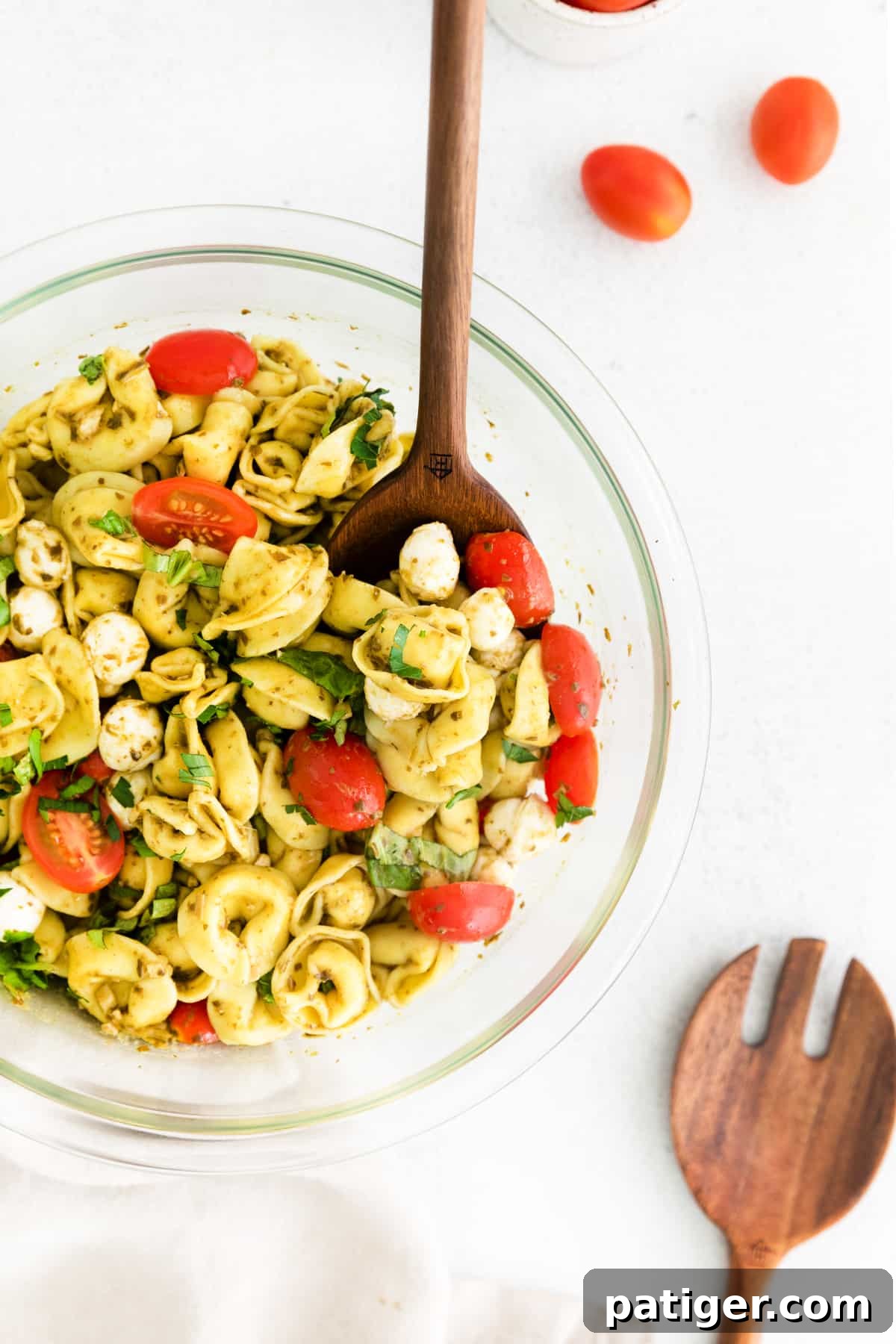 A wooden serving spoon scooping a portion of a delicious cold tortellini pasta salad with vibrant pesto dressing from a glass bowl.