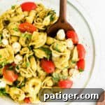 A wooden serving spoon scooping a portion of a delicious cold tortellini pasta salad with vibrant pesto dressing from a glass bowl.