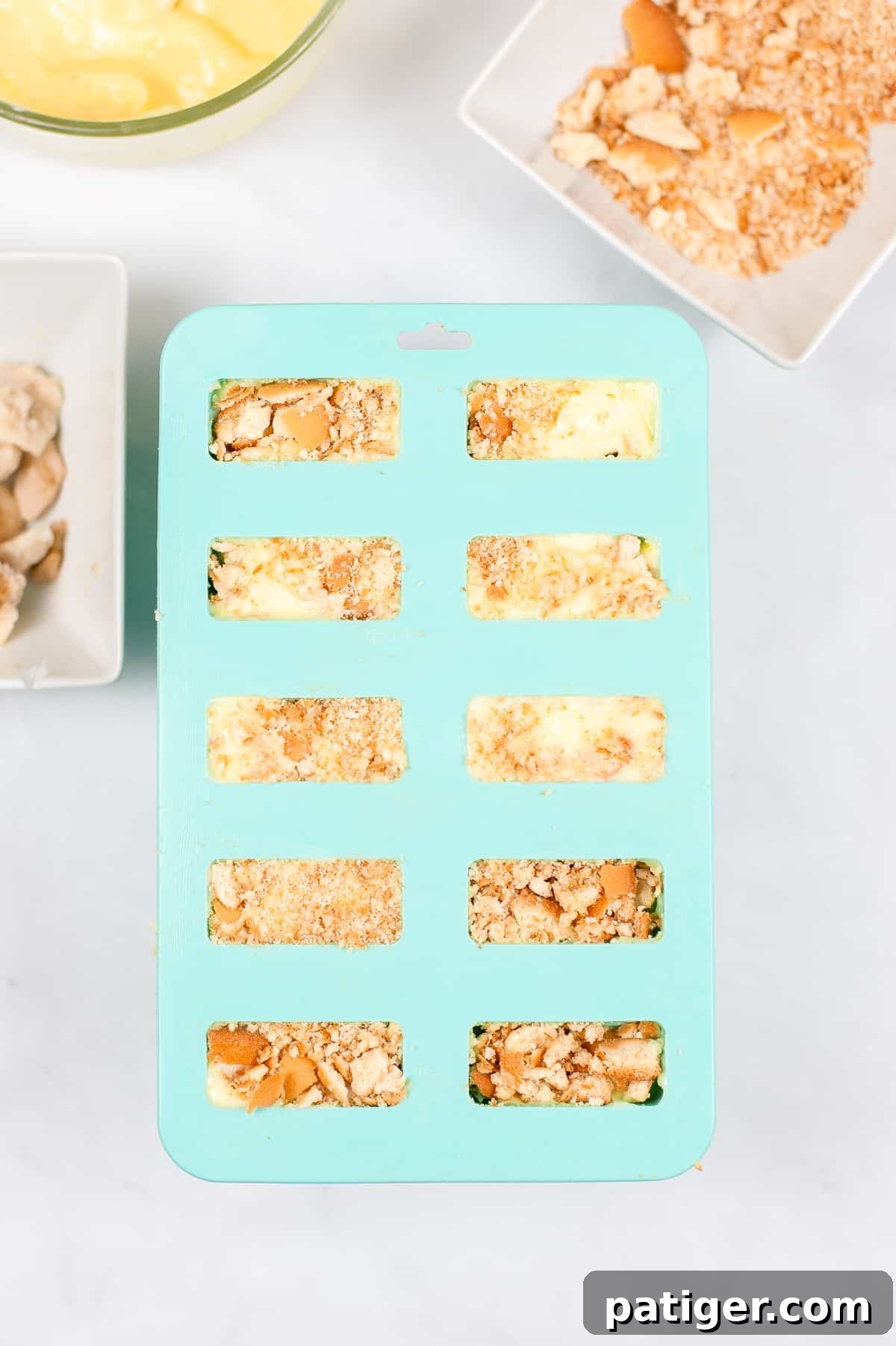 Velvet Banana Pudding Pops 4 Popsicle mold with cavities for 10 popsicles, filled with banana cream pudding, vanilla wafer crumbles, and banana slices. More ingredients are visible in the background.