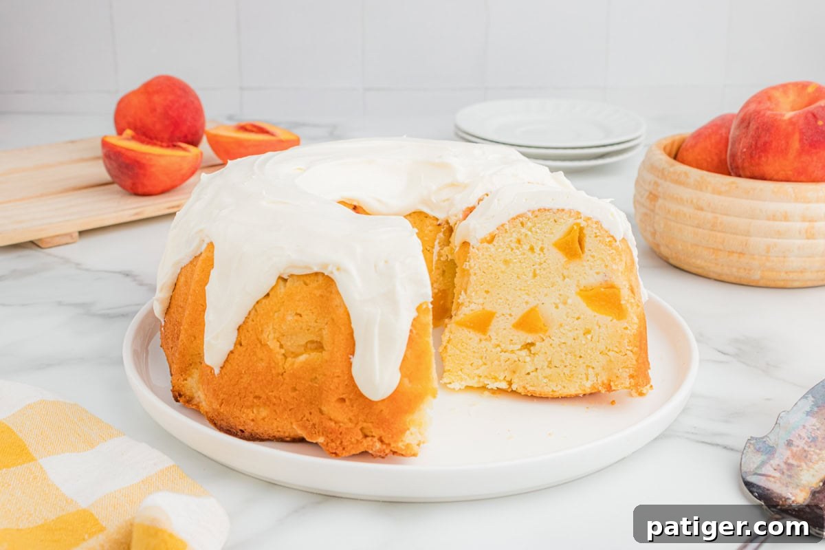 Peach bundt cake with cream cheese icing on a plate, sliced to show peach pieces inside, with fresh peaches in the background.