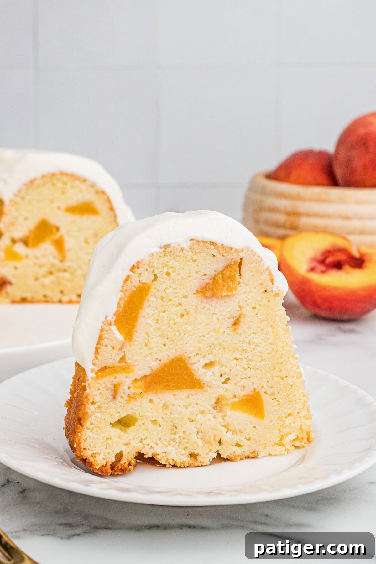 A slice of peach pound cake with cream cheese frosting on a plate with fresh peaches and rest of the cake in the background.