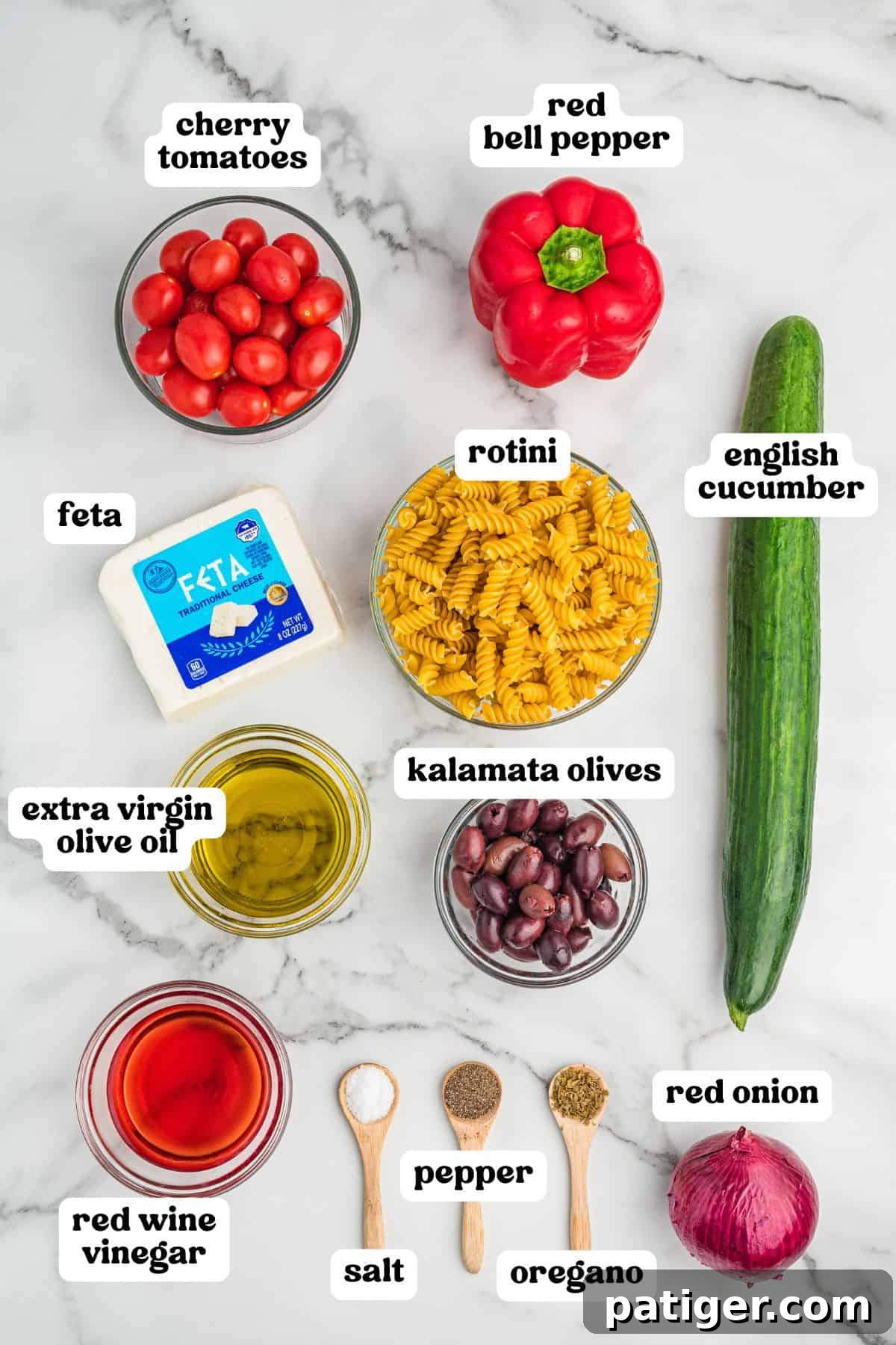 An overhead view of all the fresh ingredients artfully arranged for Greek pasta salad preparation: vibrant cherry tomatoes, crisp red bell pepper, refreshing English cucumber, sharp red onion, creamy feta cheese, curly rotini pasta, briny kalamata olives, and small bowls containing extra virgin olive oil, red wine vinegar, salt, pepper, and dried oregano.