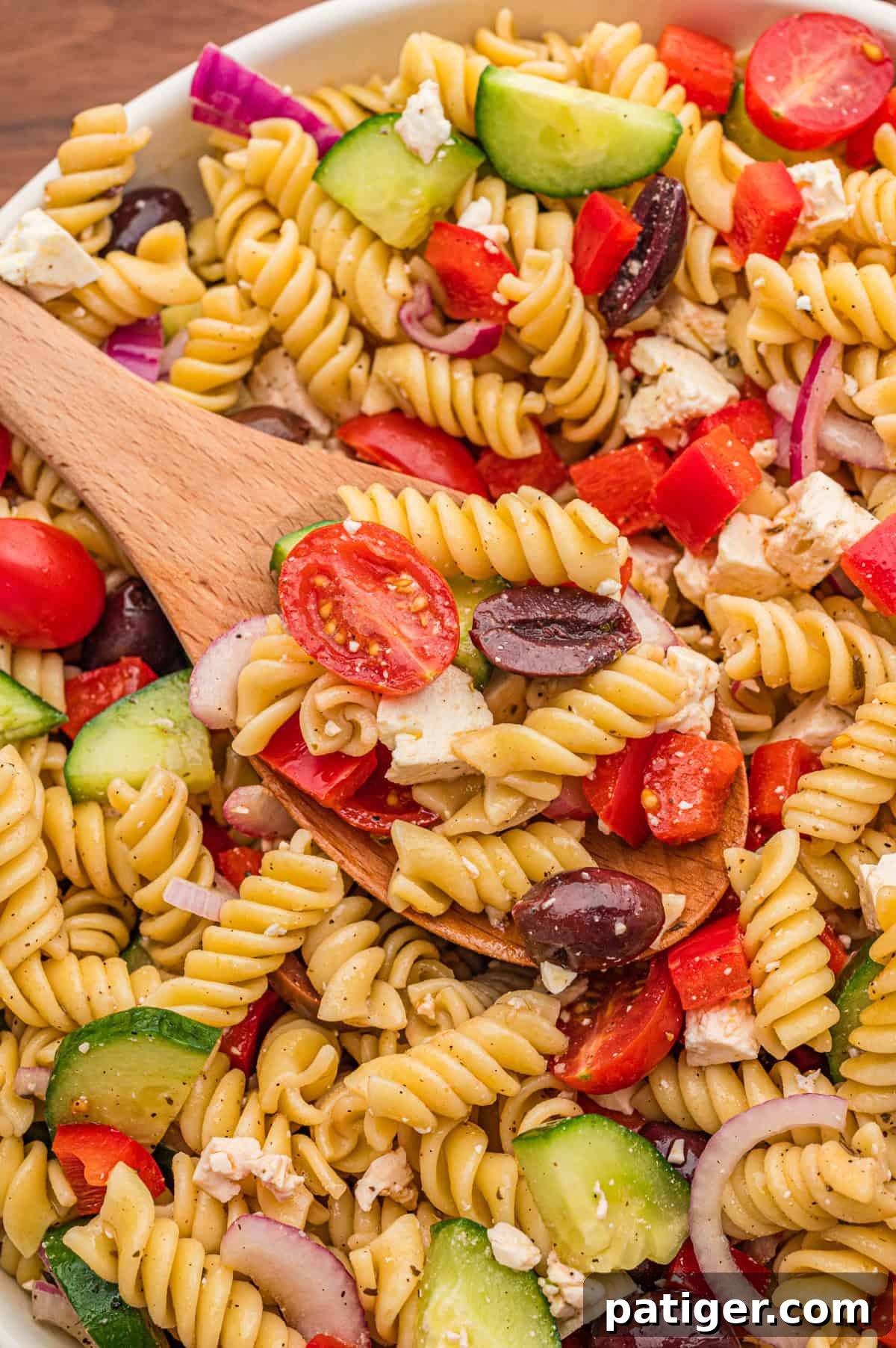 A serving spoon lifting a generous portion of colorful Greek pasta salad, beautifully showcasing the rotini pasta, halved cherry tomatoes, crisp cucumber slices, red bell pepper, briny kalamata olives, creamy crumbled feta cheese, and finely chopped red onion, all glistening with the homemade dressing.