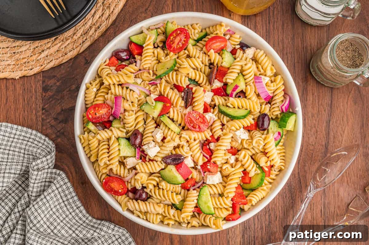 A pristine white ceramic bowl filled to the brim with vibrant Greek pasta salad, artfully placed on a rustic wooden surface. The salad showcases a rich mix of curly rotini pasta, juicy cherry tomatoes, crisp cucumber slices, dark kalamata olives, creamy crumbled feta cheese, and finely diced red onion and red bell pepper.