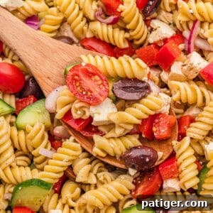 Greek pasta salad recipe with rotini, halved cherry tomatoes, cucumber slices, red bell pepper, kalamata olives, feta cheese, red onion, and homemade dressing served on a wooden spoon.