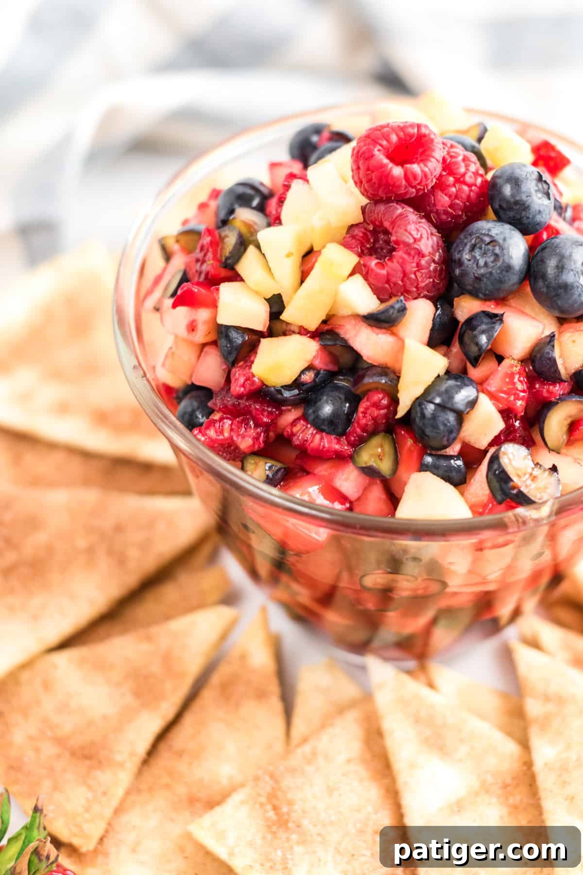 clear bowl of fruit salsa made with raspberries, strawberries, blueberries, and apples. The bowl is surrounded by cinnamon sugar tortilla chips on a platter.