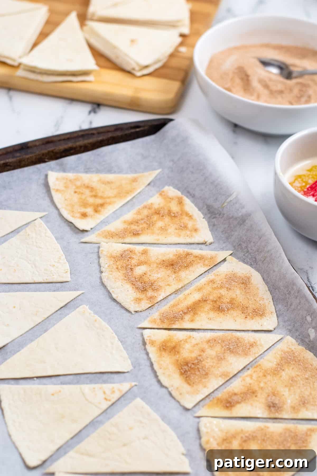 Triangular pieces of flour tortilla on parchment-lined baking sheet. Half are covered in butter, cinnamon, and sugar while the others are still plain. Bowl of cinnamon sugar and more tortilla can be seen in the background.