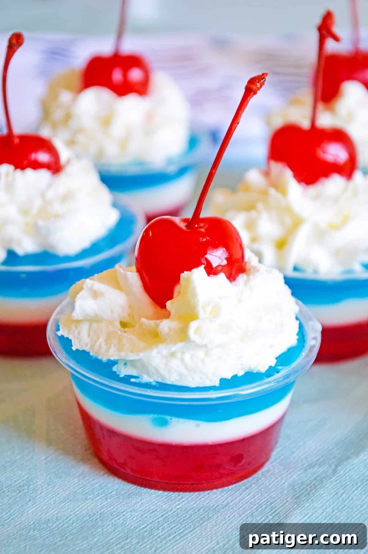 Layers of red, white, and blue gelatin topped with whipped cream and a maraschino cherry in a small clear plastic cup, perfect for a 4th of July party.