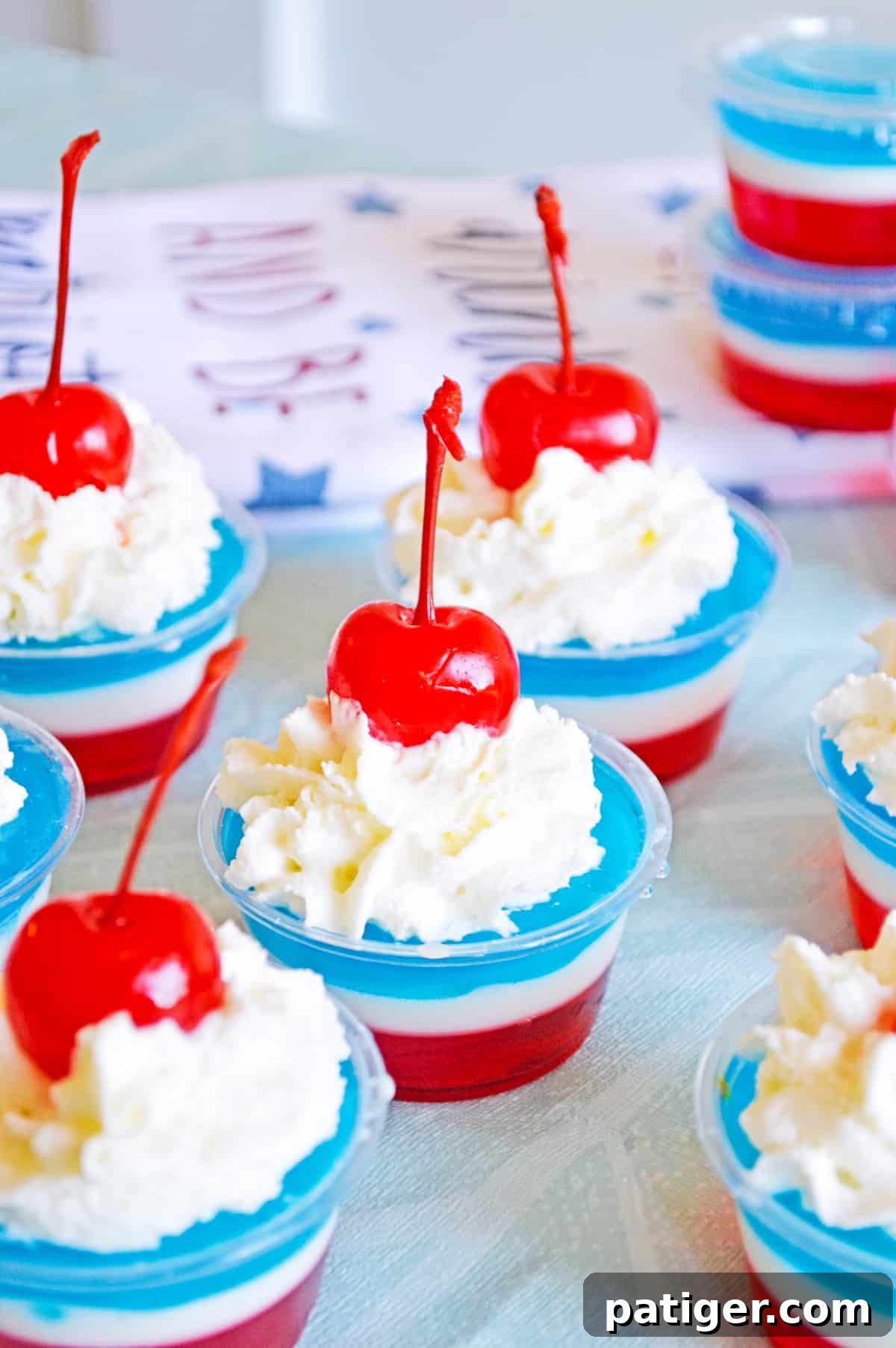 A close-up of several red, white, and blue patriotic jello shots topped with whipped cream and cherries in small disposable plastic cups.