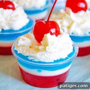 Layers of red, white, and blue gelatin topped with whipped cream and a maraschino cherry in a small clear plastic cup, illustrating the 4th of July Jello Shots.