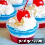 Layers of red, white, and blue gelatin topped with whipped cream and a maraschino cherry in a small clear plastic cup, showcasing the 4th of July Jello Shots.