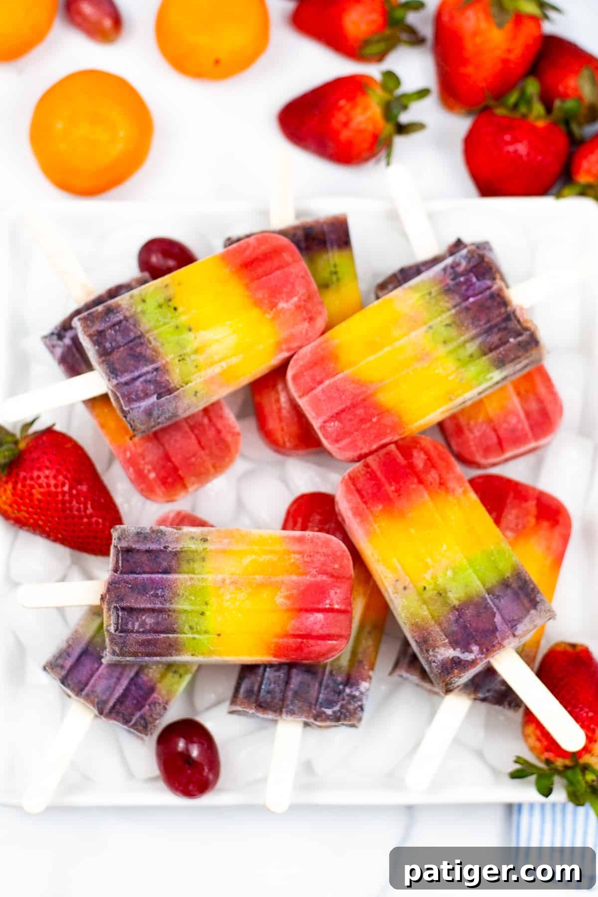 Bright and colorful rainbow fruit popsicles served on a bed of ice on a white plate. Mandarin oranges, strawberries, and grapes surround the plate, showcasing the natural ingredients and vibrant colors.