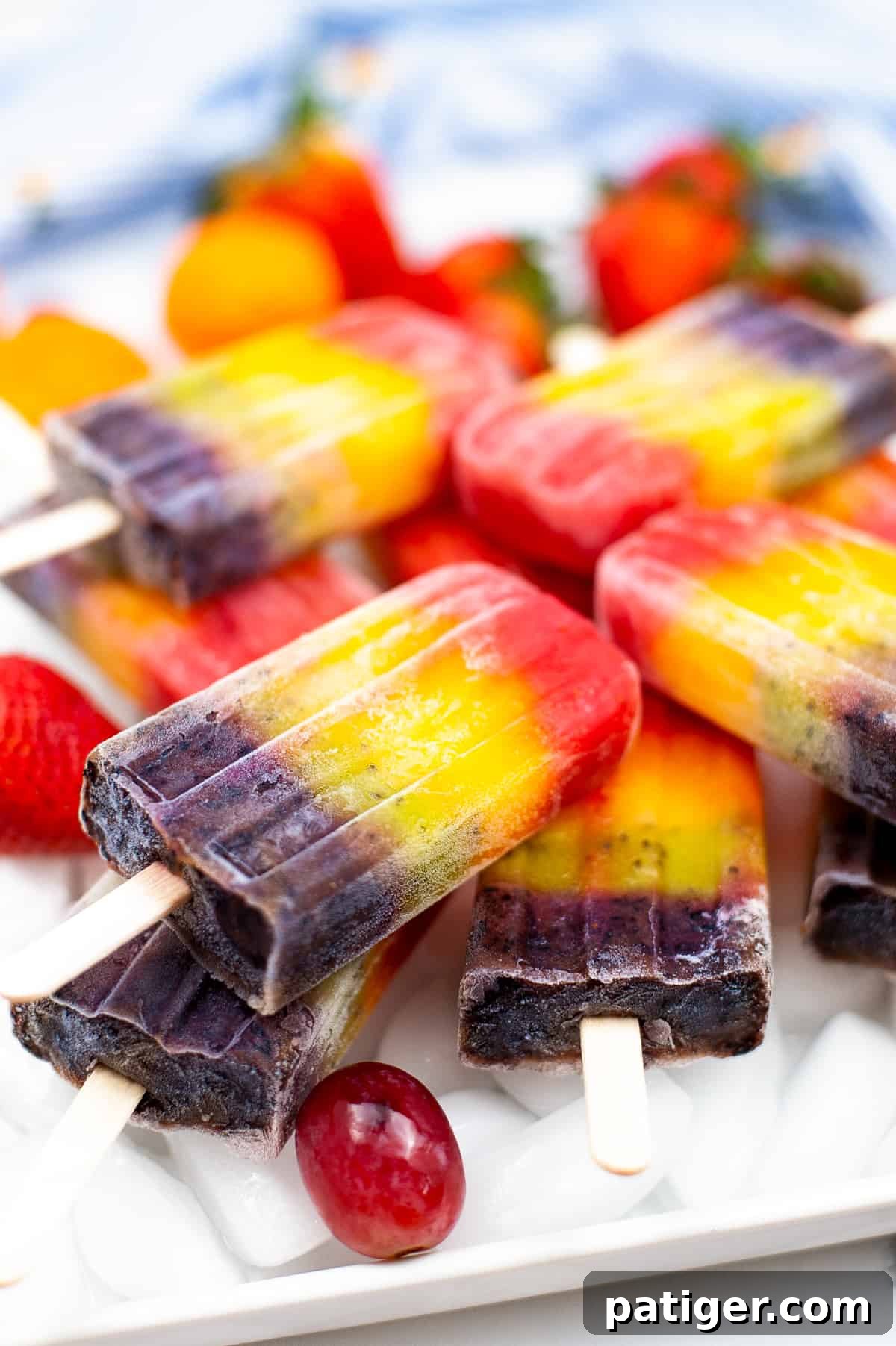 Bright and colorful rainbow fruit popsicles served on a bed of ice on a white plate, surrounded by fresh mandarin oranges, strawberries, and grapes, highlighting the natural, vibrant ingredients used.