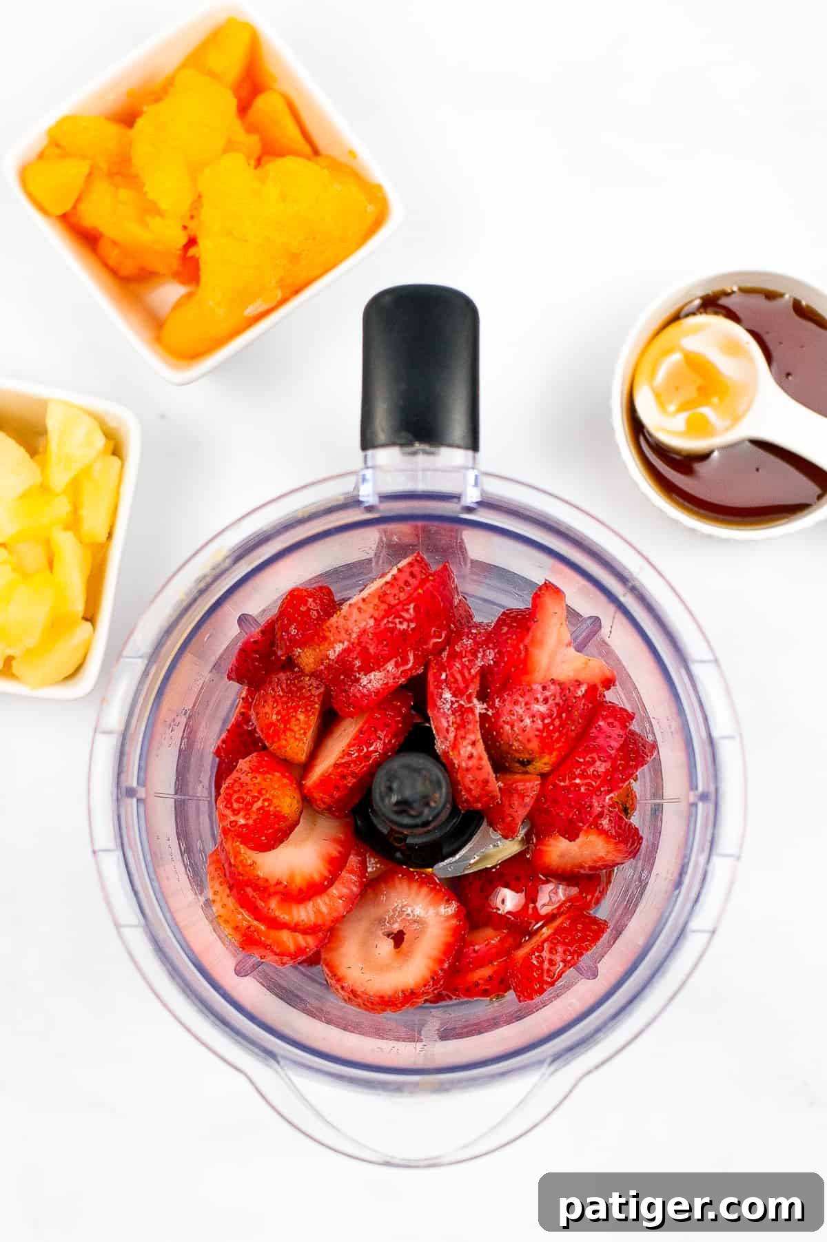 Sliced strawberries in a blender, with bowls of pineapple chunks, mandarin orange slices, and a jar of honey surrounding it, all ingredients ready for the blending process.