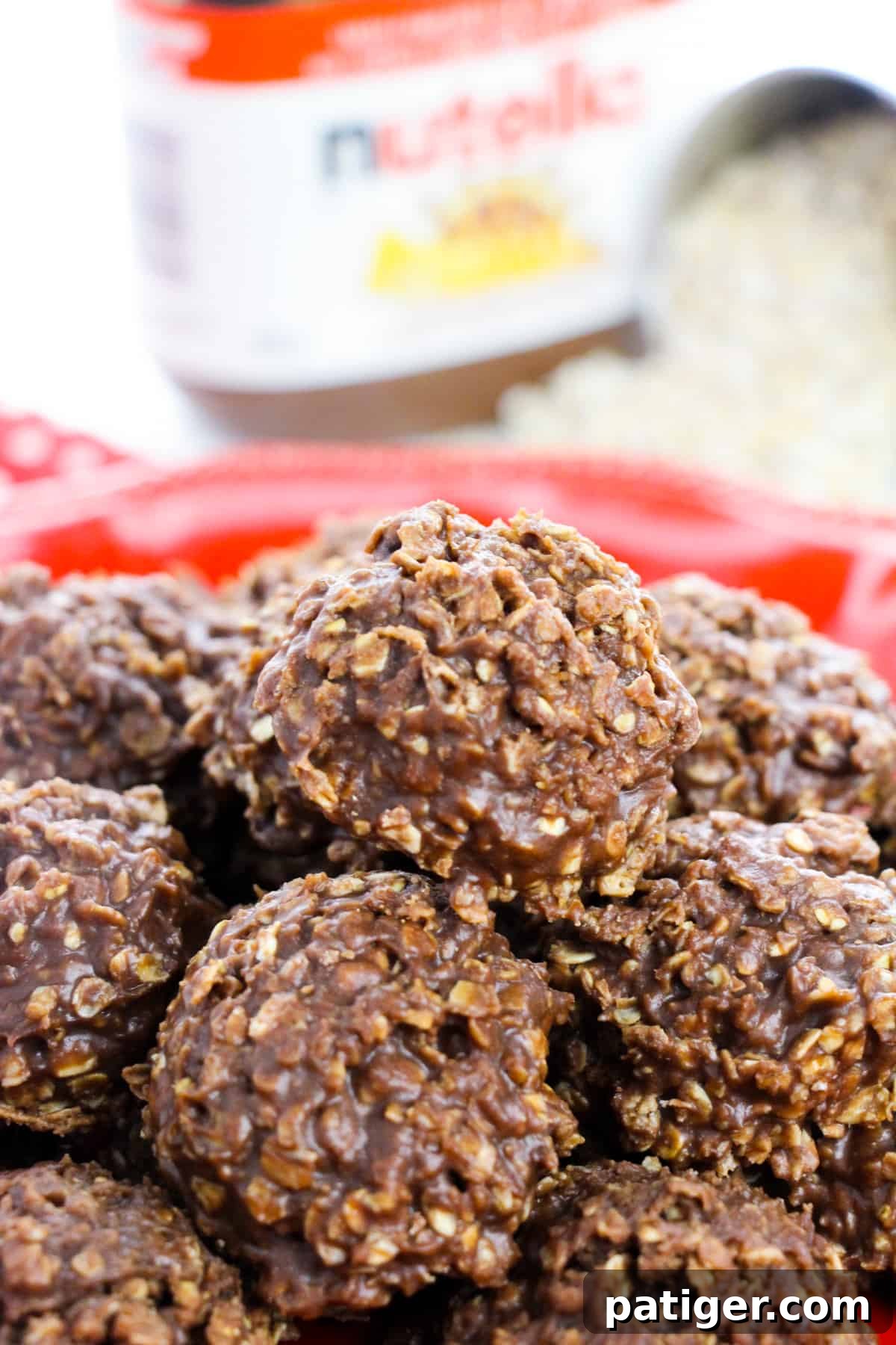 Thick chocolate nutella no bake cookies piled high on a red plate with jar of nutella and scoop of quick oats behind it