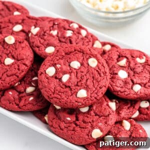 Red velvet cookies with white chocolate chips piled on white serving platter with bowl of white chocolate chips in the background