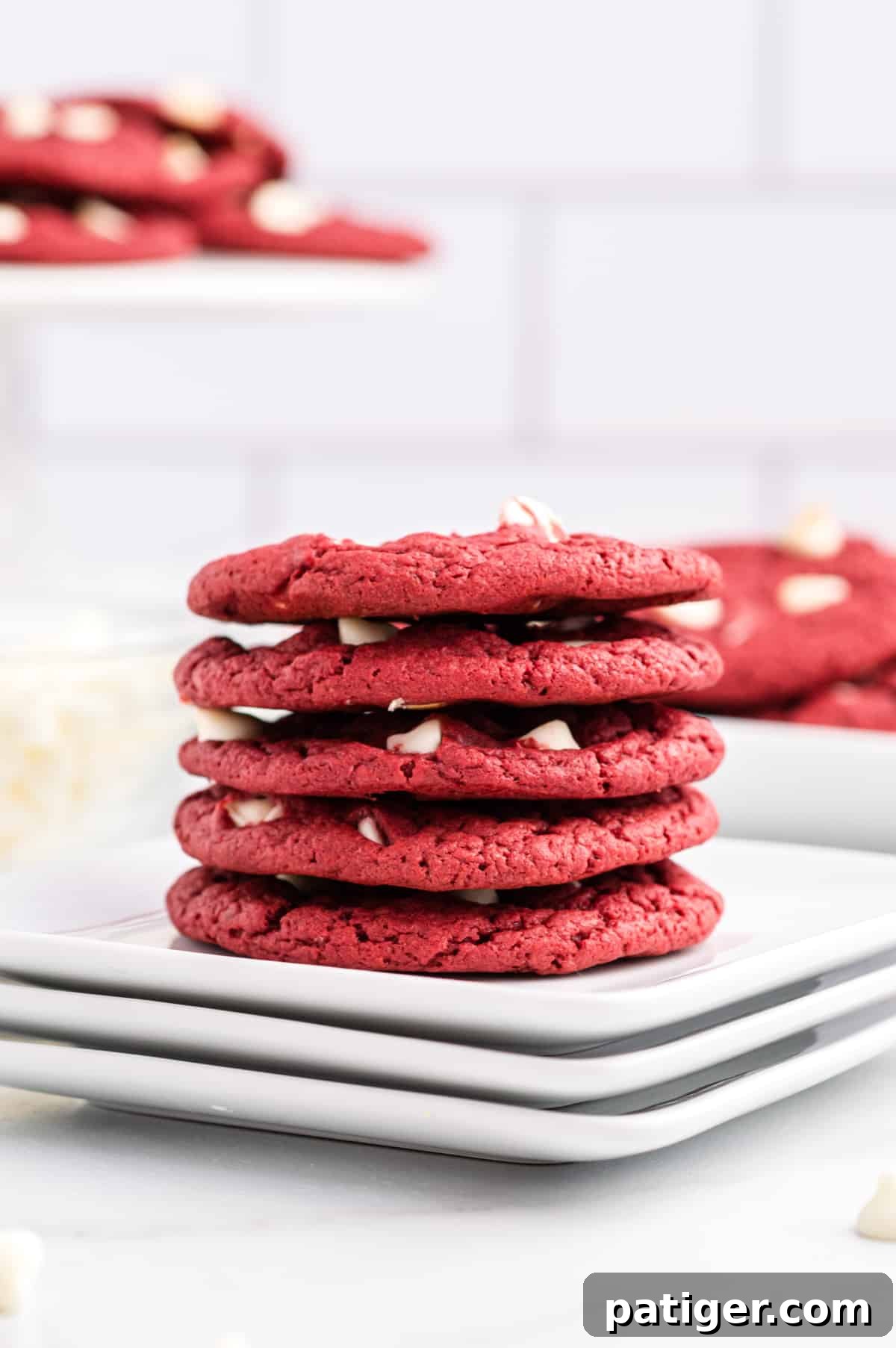 Red velvet white chocolate cookies stacked on white plates. More cookies can be seen in background.