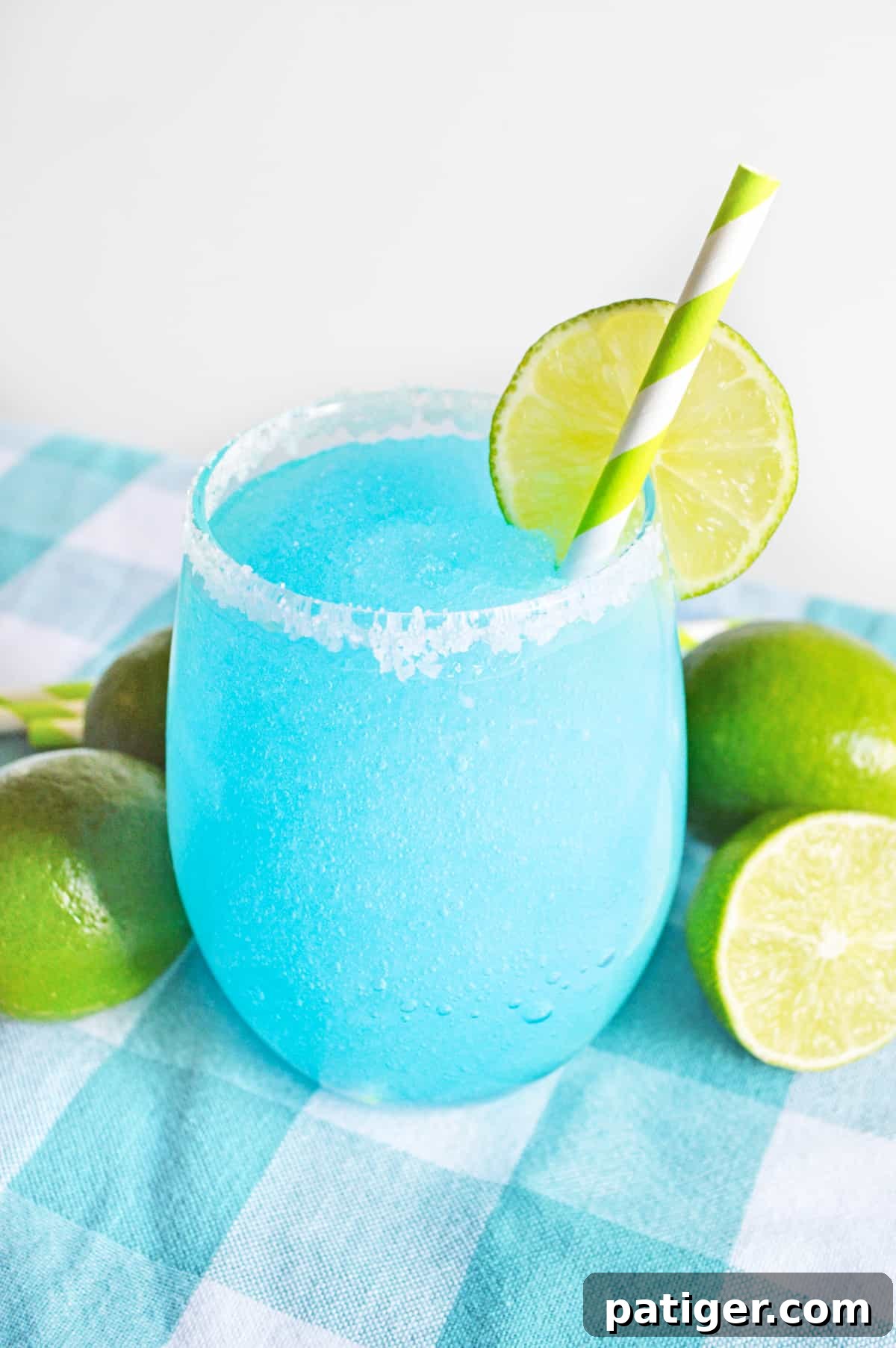 Bright blue frozen margarita garnished with lime and served in a glass rimmed with coarse sugar. Limes are next to glass on blue tablecloth.