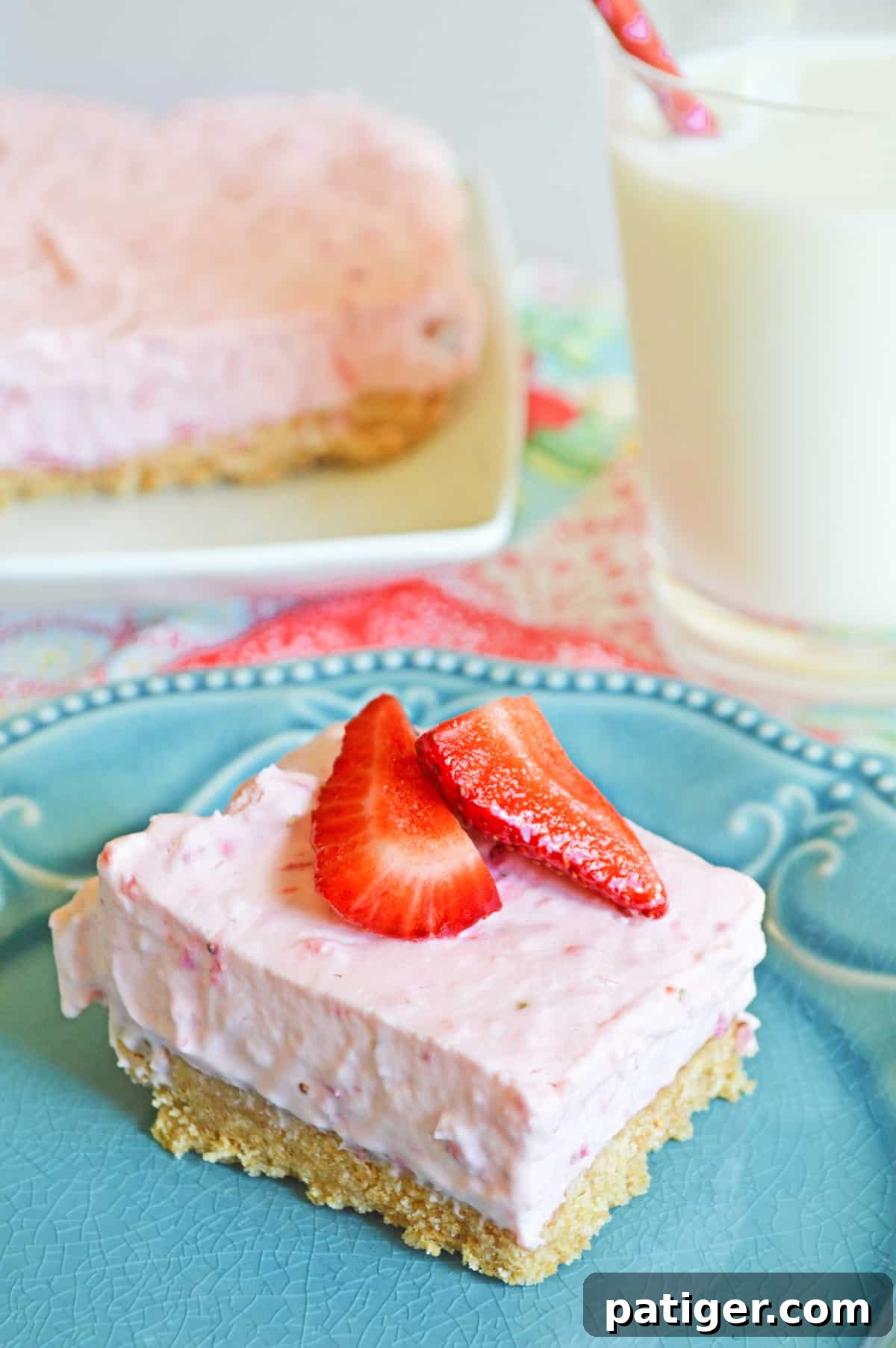 A vibrant No-Bake Strawberry Cheesecake Bar featuring a thick graham cracker crust and topped with glistening fresh strawberries, artfully presented on a blue plate. In the blurred background, a glass of milk and more delicious bars await.