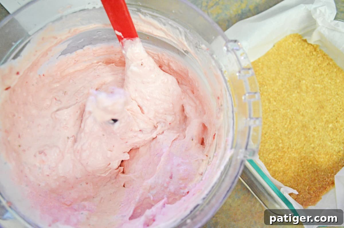 A large bowl containing the creamy strawberry cheesecake mixture alongside a square baking pan with a golden graham cracker crust, illustrating the preparation process.