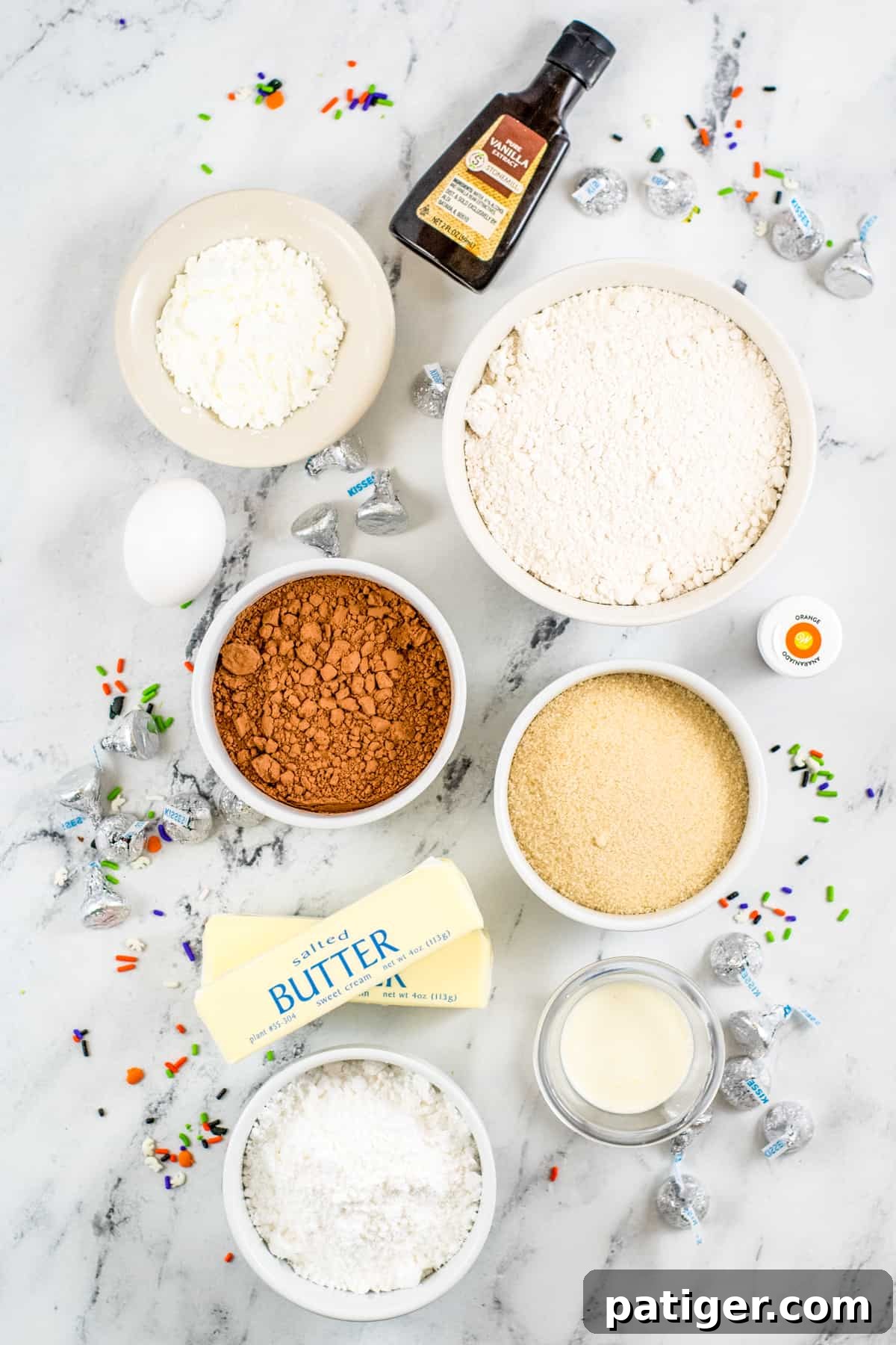 Ingredients on countertop: cornstarch, flour, cocoa powder, sugar, cream, salted butter, egg, vanilla, orange gel food coloring, powdered sugar, Hershey kisses, and sprinkles