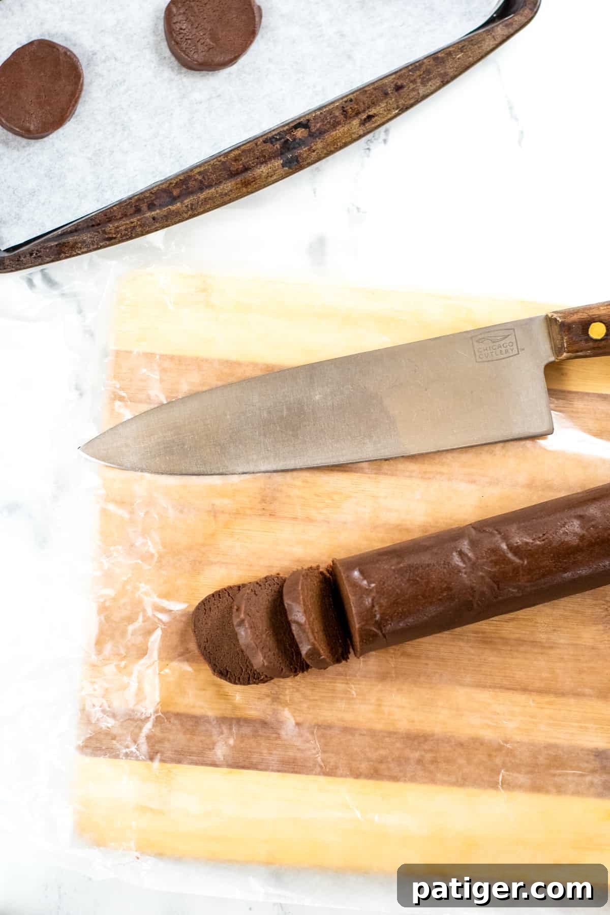 Cutting board with log of chocolate cookie dough, some of which as been cut into slices. Large knife is on cutting board and lined baking sheet with slices of cookies are shown as well.