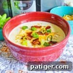 A vibrant red bowl filled with creamy crockpot loaded baked potato soup, generously topped with crispy bacon, fresh scallions, and melted cheese, ready to be enjoyed.