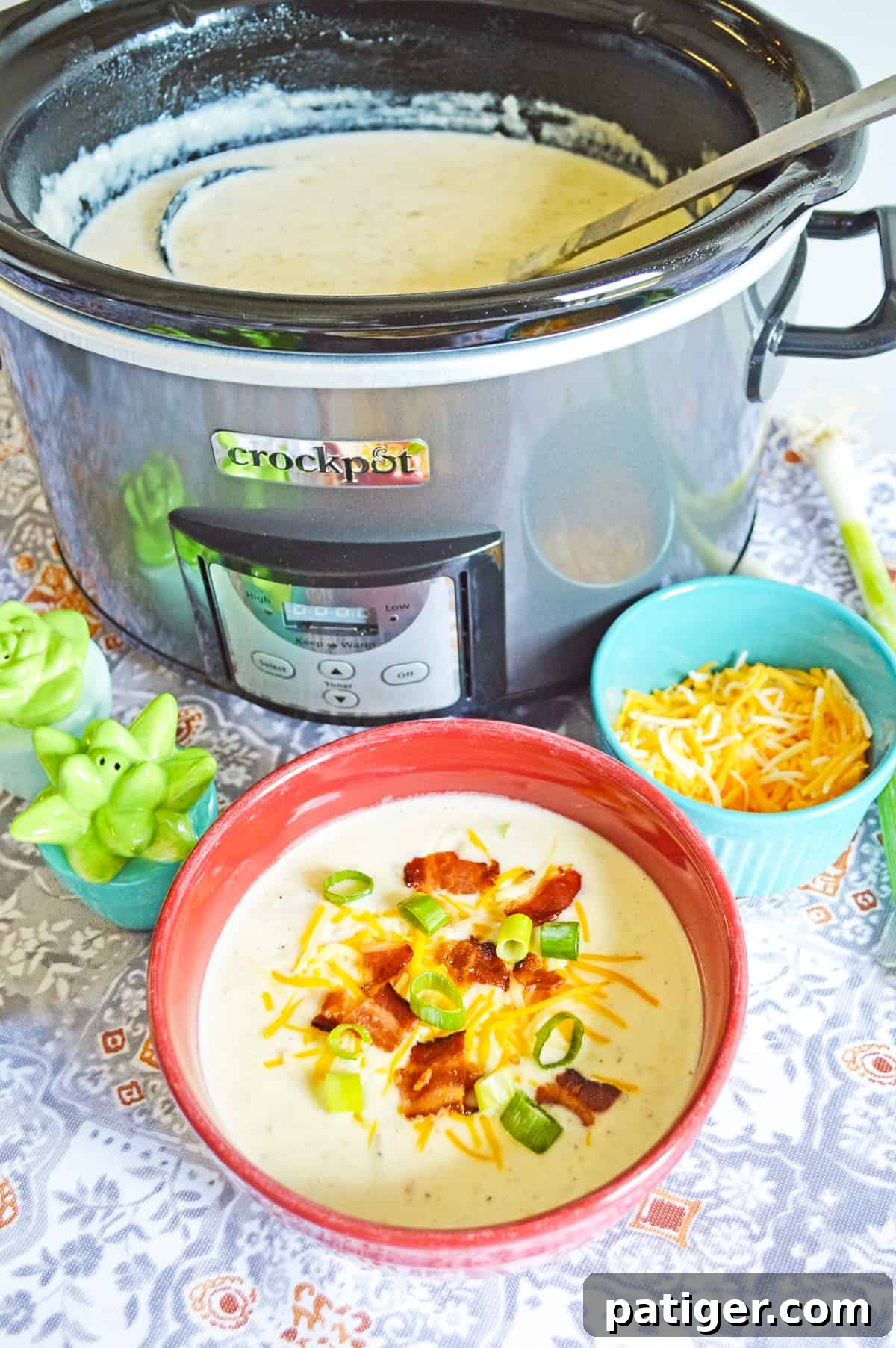 A crockpot full of simmering baked potato soup sits beside a perfectly garnished bowl of the soup, adorned with fresh scallions, cheese, and bacon. Nearby, a bowl of extra shredded cheese and salt and pepper shakers await.