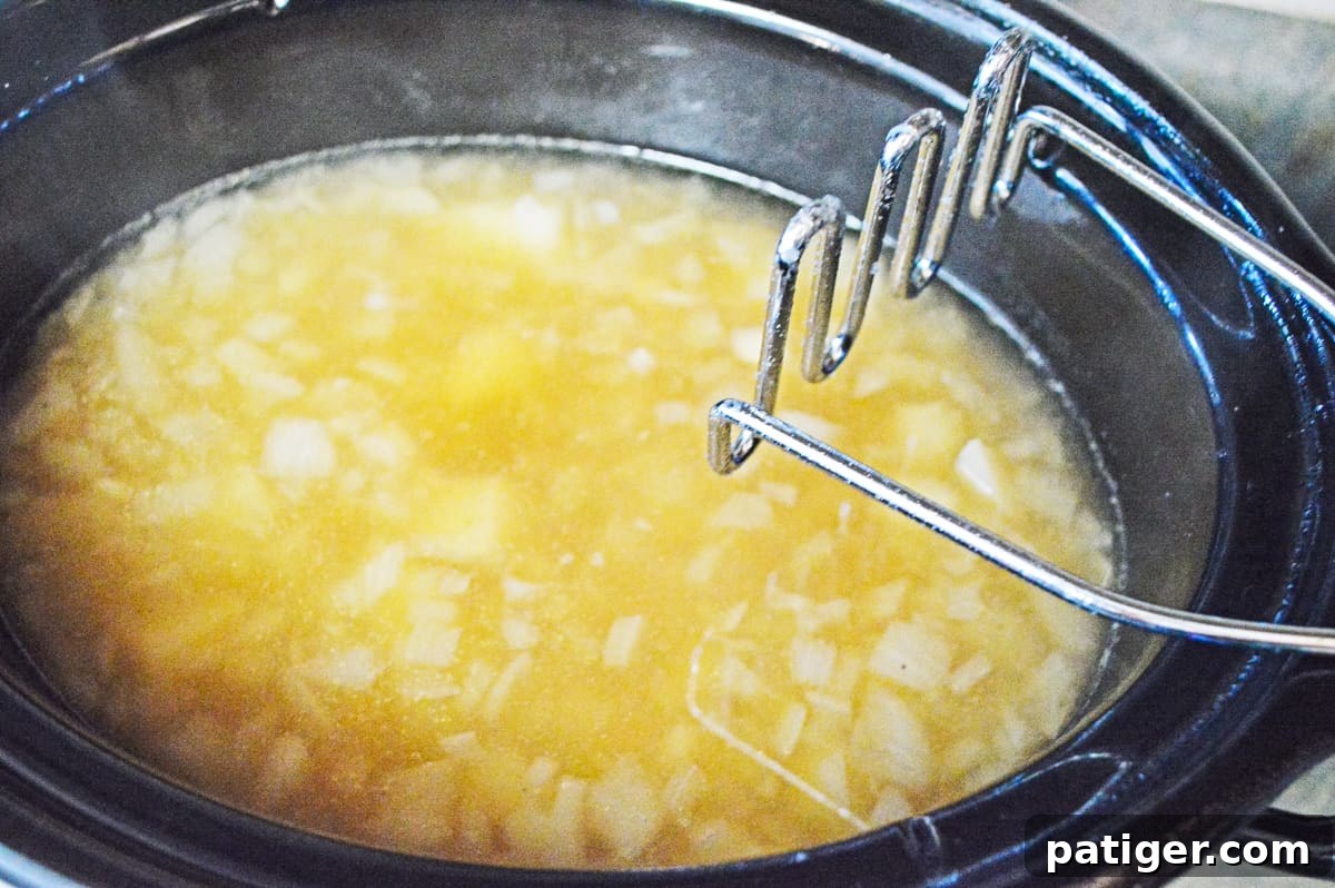A potato masher is being used to break down tender potatoes inside a crockpot, which also contains chicken broth, onions, salt, and pepper, creating a creamy soup base.