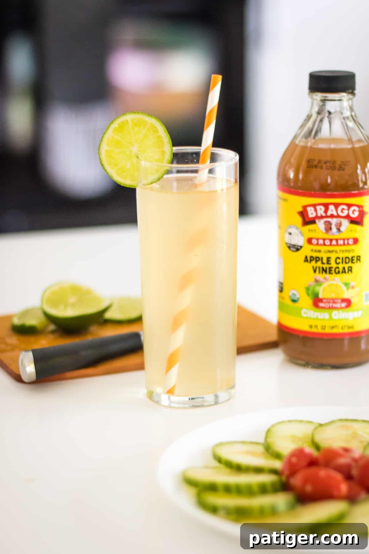 A sparkling apple cider vinegar mocktail with fresh lime slices, set against a backdrop of a cutting board with a whole lime, a cucumber salad, and a bottle of apple cider vinegar, evoking a sense of fresh and healthy living.