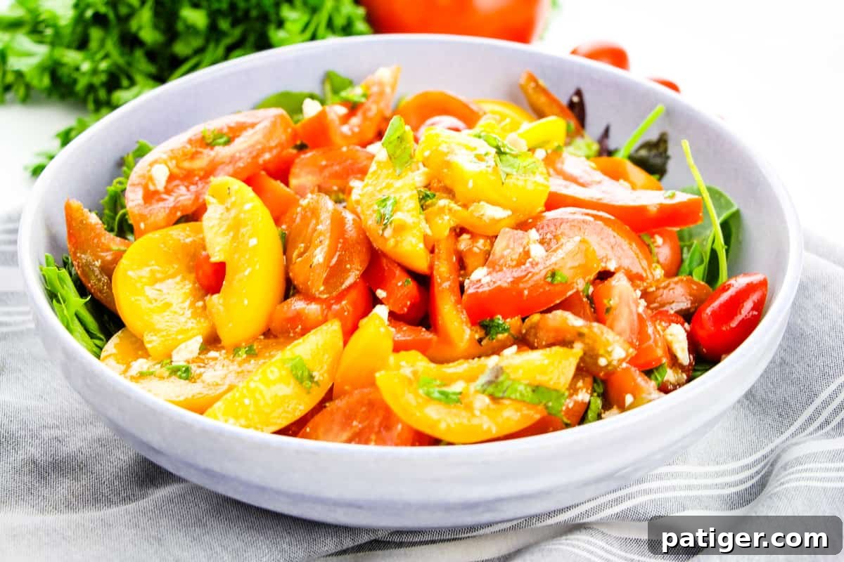 A colorful tomato salad, featuring a variety of heirloom tomatoes, crumbled feta cheese, and a glistening balsamic vinaigrette dressing, beautifully served on a bed of fresh green lettuce in a light grey bowl.
