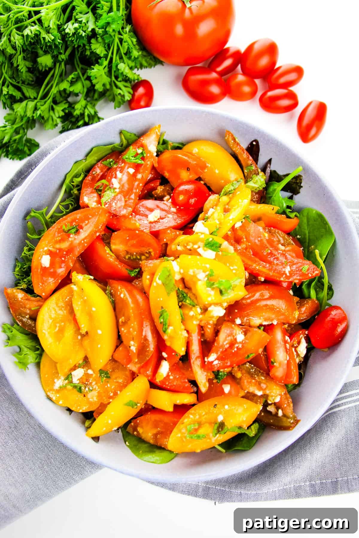 An overhead view of a vibrant heirloom tomato salad presented in a large serving bowl, elegantly placed over a bed of fresh greens. Fresh herbs and whole, colorful tomatoes are arranged artfully in the background, emphasizing the salad's fresh ingredients.