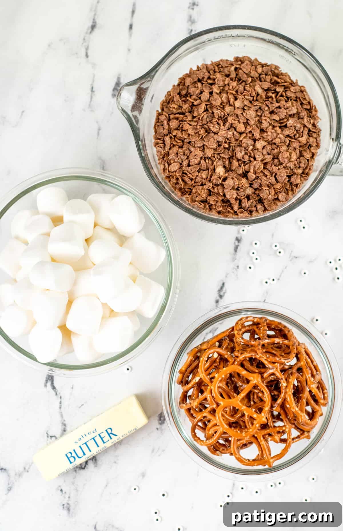 Ingredients for Rice Krispie Treat Spiders laid out: a bowl of fluffy marshmallows, a bowl of rich chocolate krispy cereal, a stick of butter, and a bowl of thin pretzels on a kitchen countertop.