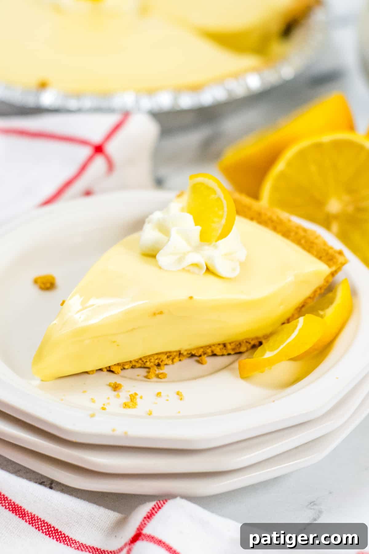 A vibrant slice of Lemon Icebox Pie on a stack of white plates, garnished with a dollop of whipped cream and a fresh lemon slice. The full pie and additional fresh lemons are visible in the soft-focus background, evoking a bright, summery feel.