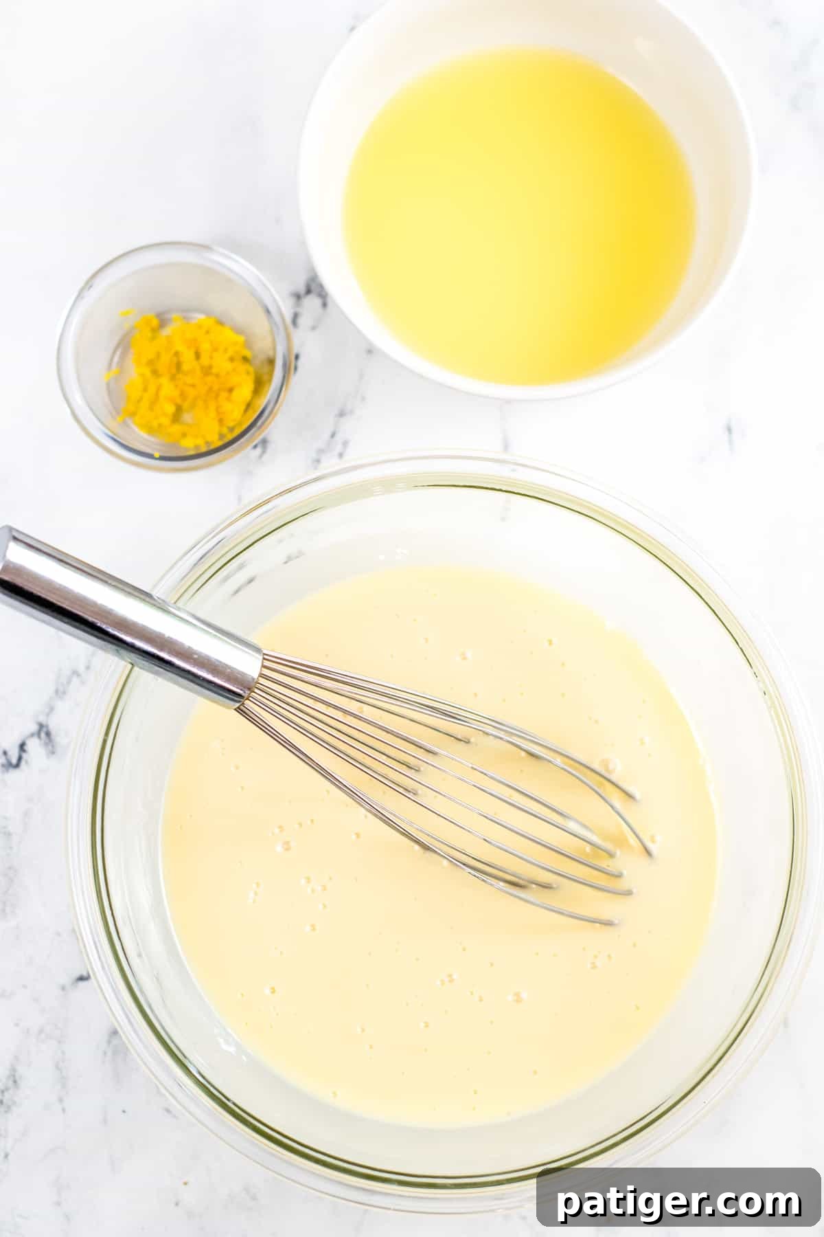 A whisk in a bowl of sweetened condensed milk, with separate bowls of lemon zest and lemon juice beside a large mixing bowl, illustrating the filling preparation process.