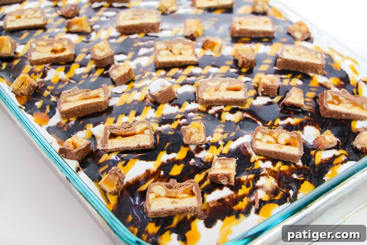 Sheet cake topped with cool whip, caramel, chocolate sauce, and snickers candy, showcasing the rich layers.