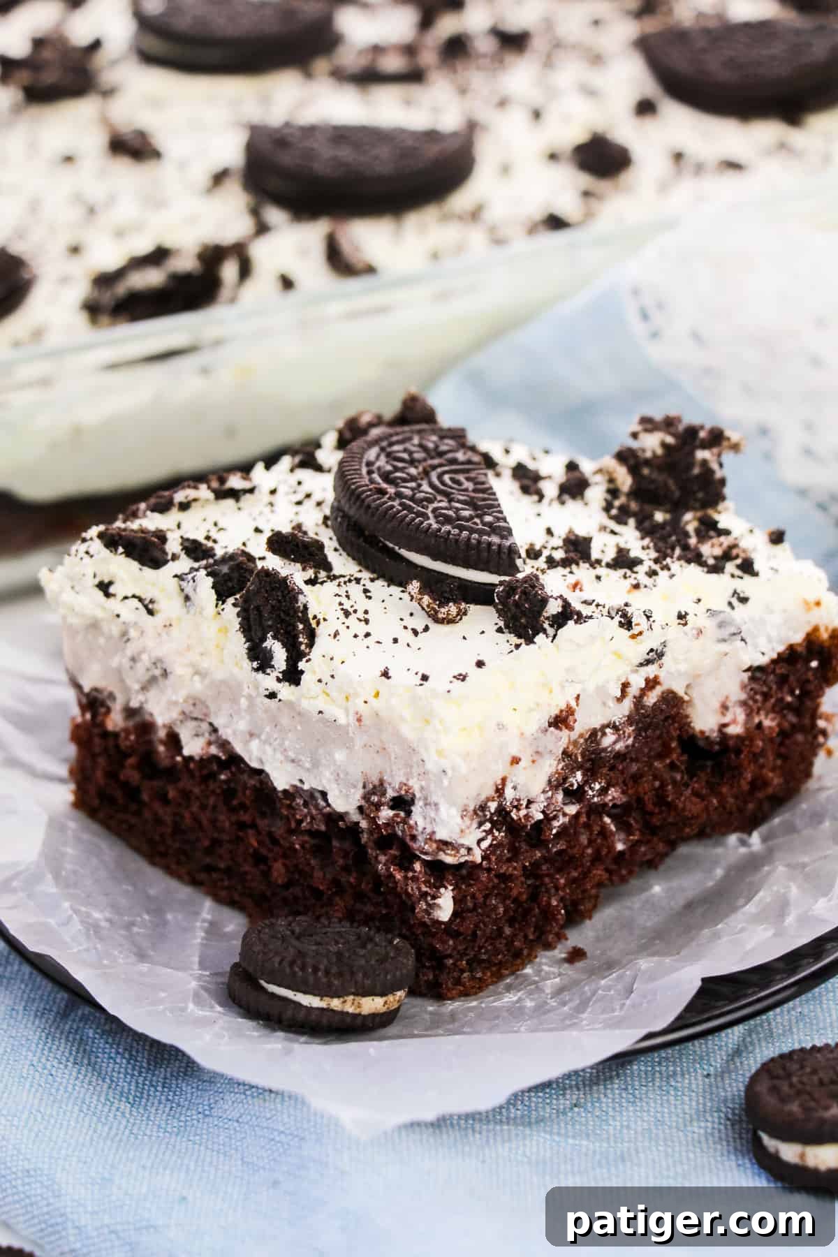 Cookies & Cream Poke Perfection 2 Square slice of OREO Poke Cake topped with Cool Whip and pieces of OREO cookies. Whole OREO cake in 9 x 13 baking dish behind plate.