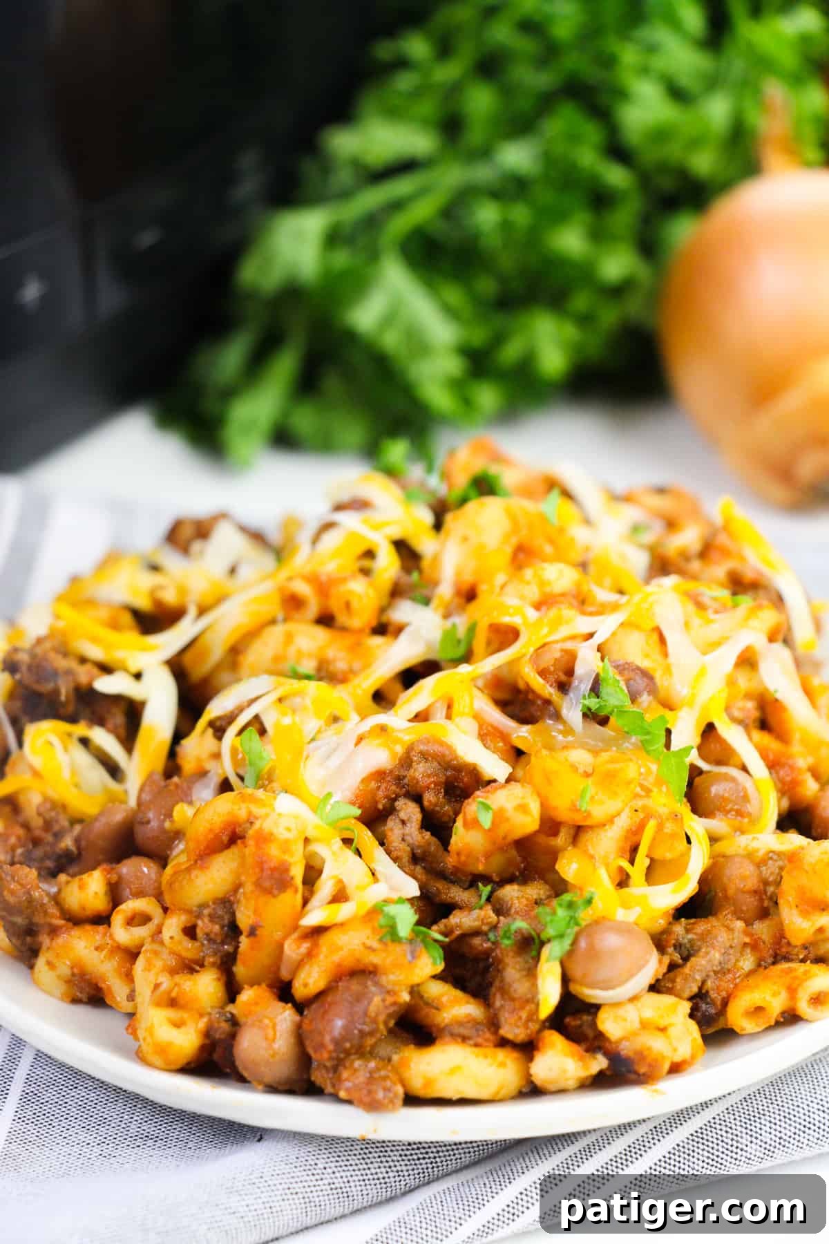 Crockpot chili mac with ground beef and shredded cheese on a white plate with fresh herbs and onion, with a slow cooker in the background.