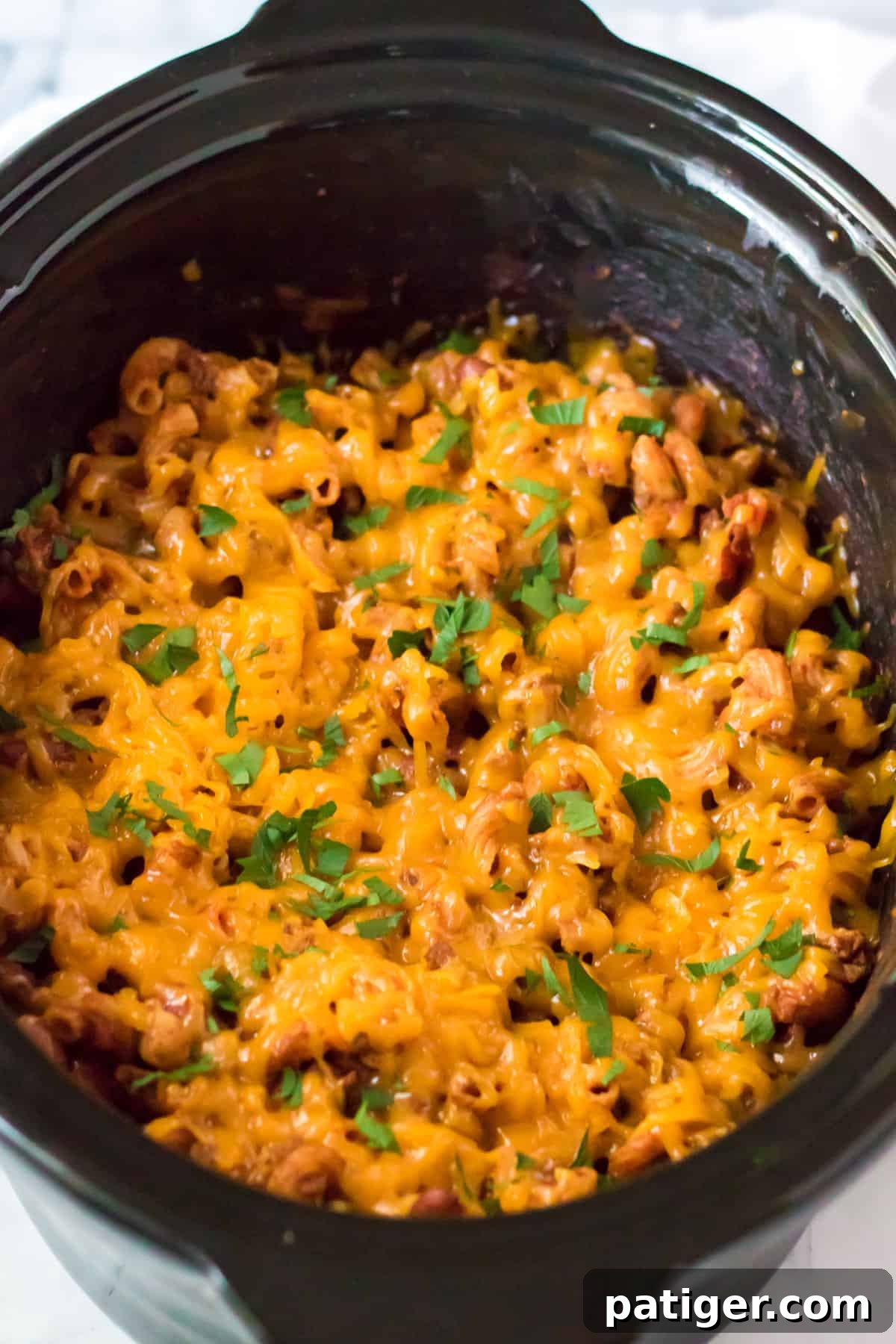 Close-up of slow cooker chili mac in a bowl, showcasing the melted cheese, beans, and garnished with chopped parsley.