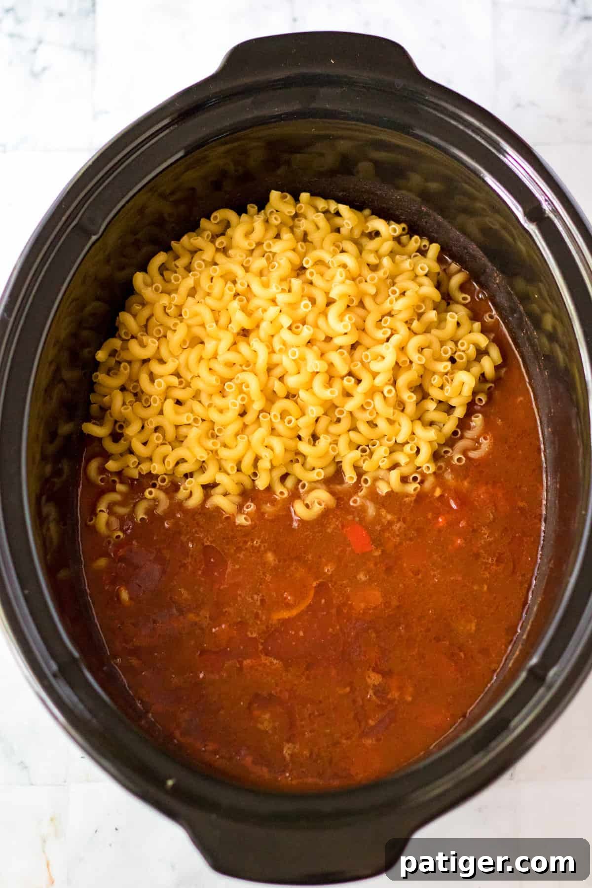 Uncooked elbow macaroni noodles being added to the chili mixture in the crock pot.