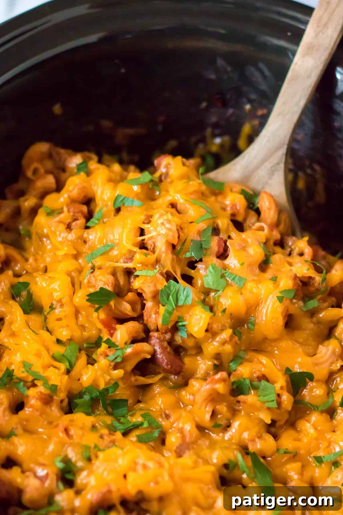 A wooden spoon scooping ground beef chili mac and cheese directly out of the slow cooker.