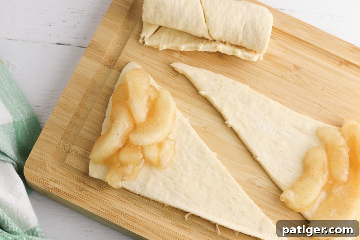 Two triangles of raw crescent roll dough on a cutting board. A spoonful of apple pie filling in on the wide end of each 