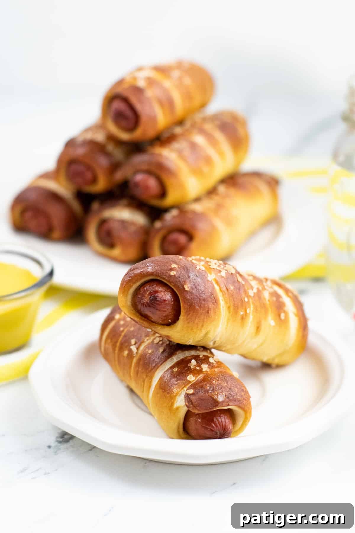 Homemade pretzel dogs served on a white plate with a bowl of mustard and more pretzel wrapped hot dogs in the background.