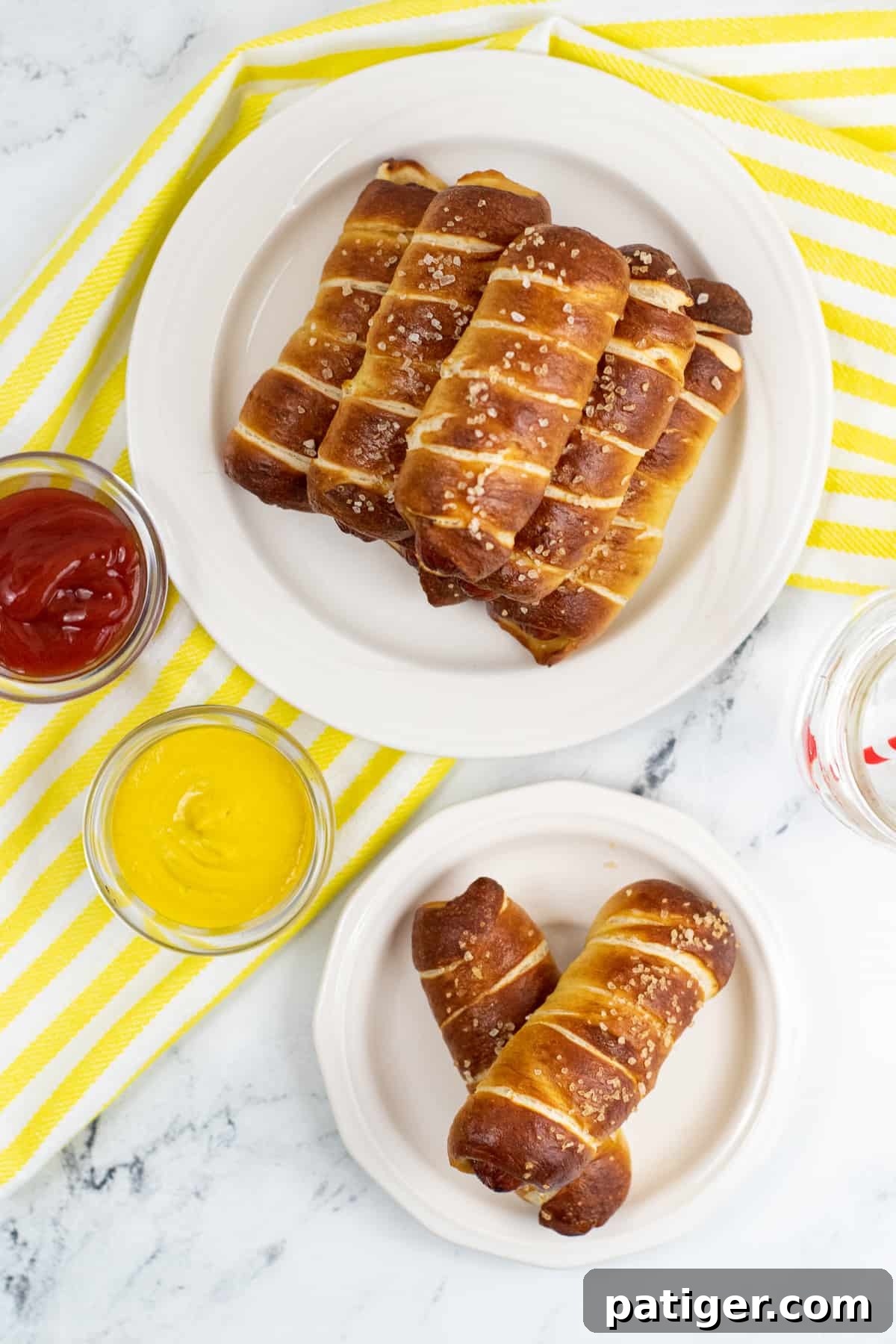 Top-down view of pretzel dogs on table both stacked on serving platter and on individual plate. Bowls of ketchup and mustard are next to them.